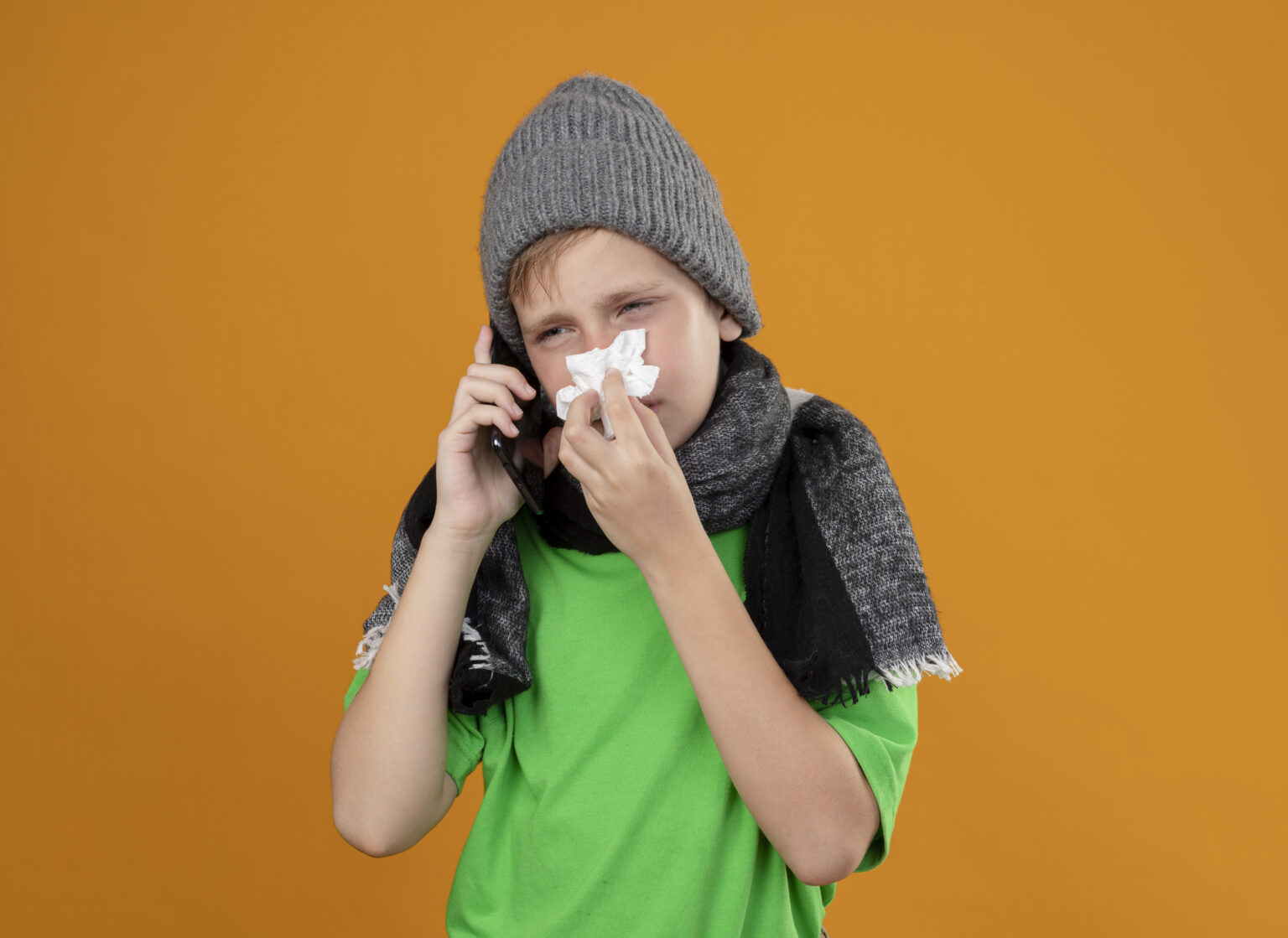 ill little boy wearing green t-shirt in warm scarf and hat feeling unwell talking on mobile phone wiping nose with paper napkin suffering from cold standing over orange background