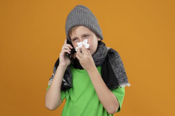 ill little boy wearing green t-shirt in warm scarf and hat feeling unwell talking on mobile phone wiping nose with paper napkin suffering from cold standing over orange background